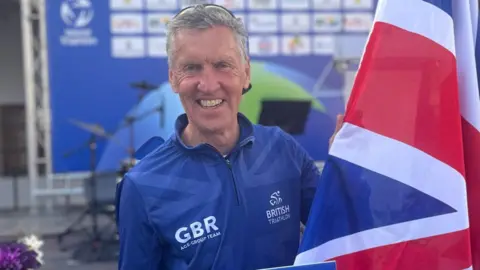 David Juggins - a man with grey hair and wearing a blue Great Britain athletics outfit - smiles as he holds a union flag after finishing an event.