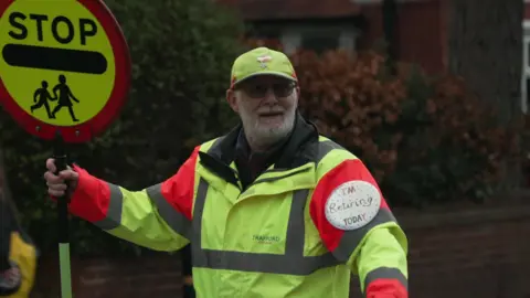 BBC Lollipop man John Astley holds up a lollipop sign bearing the words 'stop' with a symbol of children crossing a road. He is smiling and wearing a high-vis jacket and hat while stood by a crossing. 