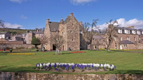 WF Millar An imposing old stone building viewed across lawns with a nice flower bed of purple and white in the foreground. There are rows of houses to the right and behind the main building.