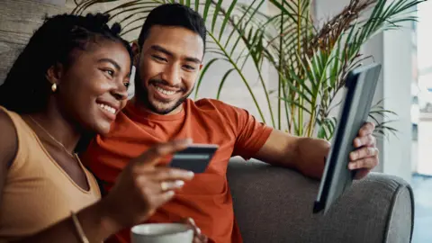 Getty Images Couple sitting on a sofa with a woman holding a bank card and a man holding a tablet computer. Both are smiling.