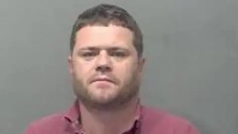 Bedfordshire Police Darren Campbell; a police custody image of a man with brown hair and a brown beard wearing a pink open-neck top.
