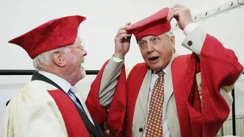 Matthew Lewis/Getty Images Sir David Attenborough, right, and his late brother Richard Attenborough. The pair are pictured joking around, wearing red gowns and mortarboard caps.