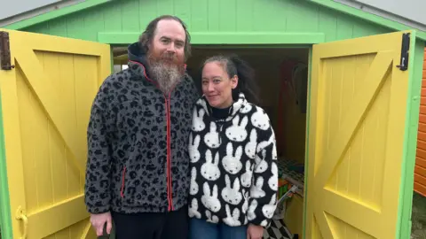 Clare Worden/BBC James Goodman Stevens and Antonia Hayes stand in front of their beach hut which is a vibrant yellow and green. James has a long beard and dark hair and is wearing a grey fleece top. Antonia has long dark hair and is wearing a black and white fleece top. 