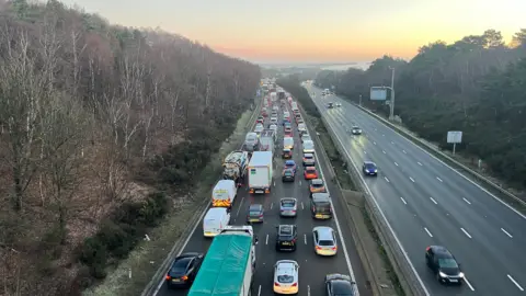 A motorway with four lanes in each direction, seen from above. One side is jammed with cars and lorries while the other has few cars, which are moving. The sky is orange as the sun rises.