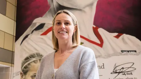 John Owen A woman with blond hair smiling into the camera in front of a mural of herself wearing an England football shirt. She is wearing a grey cardigan with a white top underneath. On the mural of herself behind her there is a signature of Kelly Smith.