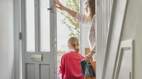 Getty Images A young child, dressed in a red school uniform cardigan is heading out of her home through the front door with her mother. We can only see the back of her head. The decor is off white and the mum is wearing a striped red and white jumper and a black handbag 