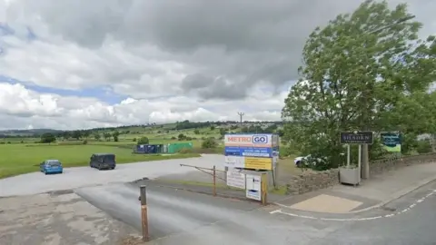 The entrance to a gravel car park, against the backdrop of some green hills