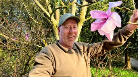 Gary Long is smiling and holding a branch down which has a huge pink magnolia flower on it. He is wearing a Trewithen branded cap and sweatshirt. 