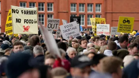 Farmers stage a protest in Cardiff Bay, with placards saying "no farmers, no food". Behind the crowd of people the red brick of Cardiff Pierhead building can be seen. 