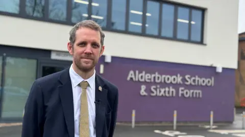 Headteacher Tom Beveridge stands in front of Alderbrook School, a modern purple and white building can be see behind him. He is wearing a blue suit, white shirt and yellow tie.