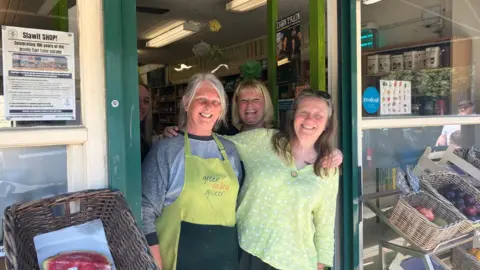 Four woman stand at the shop door of a greengrocers. They are smiling at the camera. 