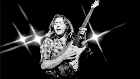 Brian Cooke/Getty Images Rory Gallagher, with long hair and checked shirt, playing a guitar