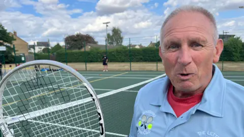 Peter Bibb, 66, has very short grey spikey hair, he is wearing a sky blue t-shirt type top with open unbuttoned collar beneath that is a red t-shirt. He is holding a tennis racquet and has a tennis court behind him.