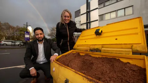 A man with dark hair and a beard is kneeling beside a full grit bin which is yellow and full of brown rock salt. he is wearing a white shirt and black suit. Beside him a woman with blond hair, wearing a dark coat, is stood smiling at the camera.