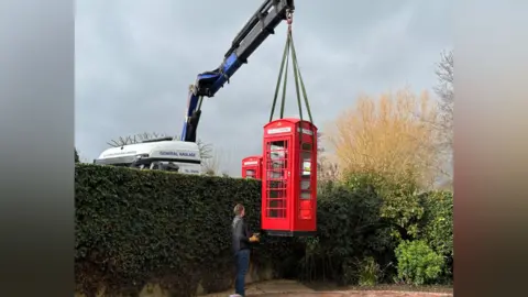 Greg Horsford A red phone box being craned into a garden over an ivy clad wall. The delivery driver is standing beside the phone box looking up at it as it dangles in mid air. Behind the wall is a lorry cab roof and another red phone box can just be glimpsed. 