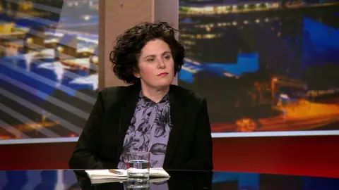 Claire Hanna, a woman with short curly dark hair, in a black blazer with a patterned purple and black blouse, sat at a black desk with a notebook and a water glass in front of her, sat in the View studio