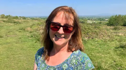 Rachel Tobin standing on Castlemorton Common, a green hill, in the sunshine, wearing sunglasses and smilling. She has shoulder-length brown hair and is wearing a summer top. The hilly Malvern landscape is behind her.