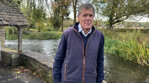 Gilbert Chalk, an older man who is wearing a long-sleeved navy top, a navy gilet with brown piping, and a white shirt underneath, stands in front of a short Cotswold stone wall, bordering a river. On the opposite bank, there are reeds and trees, and farmland on a hill in the distance.