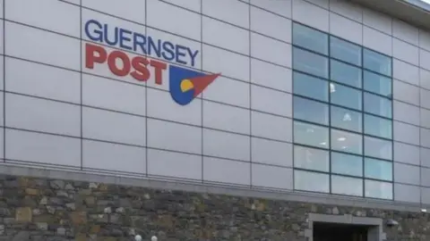 A Guernsey Post building with the company's emblem on the side of a grey building with 21 rectangle windows stacked in a cluster.