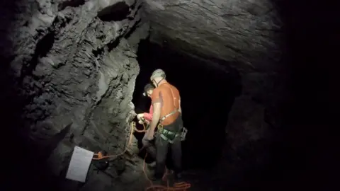 BBC Cymru Wales | Darlun The image shows two individuals wearing helmets and harnesses inside a dark cave. The man in the foreground has an orange t-shirt and green harness on and is wearing a caving helmet. In front of him, another man in red is examining rope in his hands. A piece of paper or sign is attached to the rock wall on the left side. The cave walls are rugged and textured, carved dark coloured rock in the slate mine. The scene is lit by the helmet torches being worn by the two men.