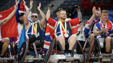 Steve Hooper is seated in the middle of a group of athletes in sports wheelchairs. They are wearing UK kit and draped in Union flags. They are all wearing silver medals on yellow ribbons and holding their arms up in a cheering gesture