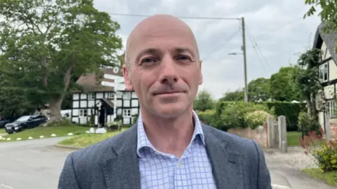A bald man in a blue jacket and blue checked shirt with black and white timbered buildings behind him