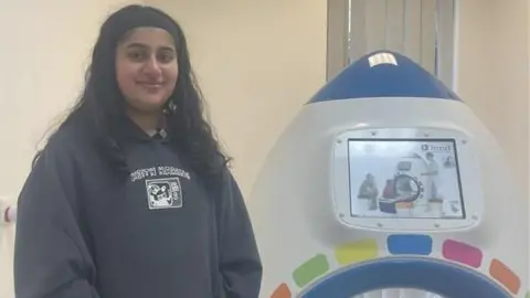 BBC Manchester Royal patient, Manha is standing in front of the new rocket scanner device, she has long dark and is wearing a grey hoodie. 