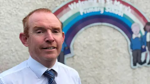 A man stands in front of a sign. The background is slightly blurred, but you can see the sign is a rainbow with children depicted either end with the words 'welcome to St Mary's Primary School' printed on top. The man has red hair and a smile. He's wearing a white shirt and a stripy tie in shades of blue. 