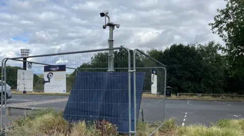 George Carden/BBC A camera pointed towards the entrance of the car park. It has fencing around it and solar panels at the front