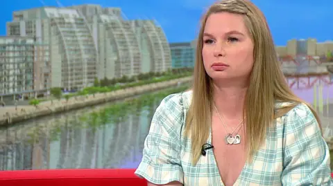 Katie Brett has long blonde hair and a pale yellow and green checked dress.