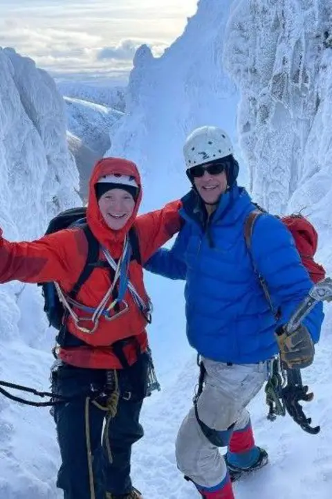 Kym McGillicuddy Two people smile on a snowy mountain with climbing gear, the one on the left wears a red jacket and the one on the right wears a blue jacket