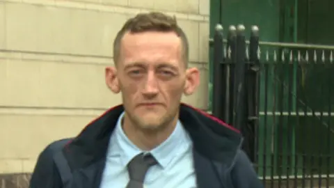 BBC Craig Rowland - Still image of a man with short brown hair wearing a light blue collared shirt, a slim gray tie and an open dark jacket. He walks in front of a tall, black gate and a Portland stone wall can be seen in the background