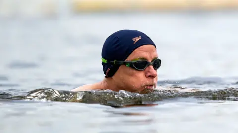 A man in goggles and a swim cap, with his head above the surface, swimming in open water