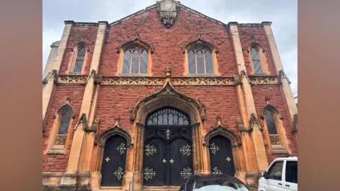 The front of a church which has red brick and one storey with arches windows and an arched doorway, with black doors and gold ornate patters on the hinges and handles.