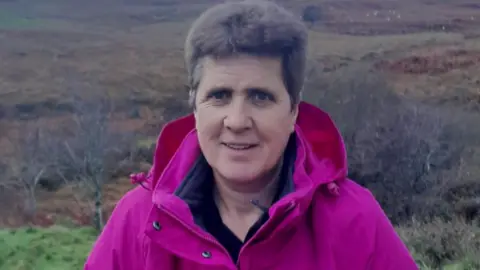 BBC A woman with short grey hair and a bright fuschia waterproof jacket stands outdoors in front of a stretch of wild Scottish countryside