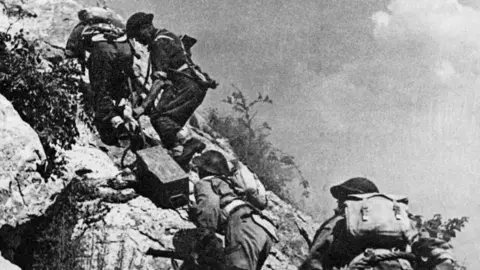 Getty A black and white image of 2nd Polish Corps soldiers carrying supplies of ammunition up a steep rocky hill during the last battle of Monte Cassino. There are four men in single file on the near-vertical slope, with packs on their backs and the second man is pulling a box. 
