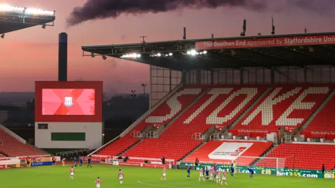 @welloffside/@simonstacpoole A stand at a football stadium, with the word Stoke picked out in white, among rows of red seats. There are players on the pitch, and smoke coming from a chimney outside the ground