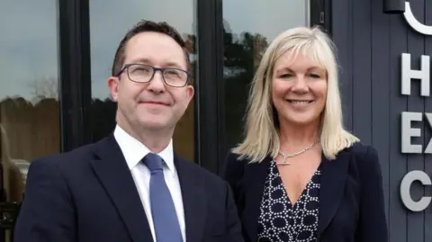 BBC A man and a woman are looking into the camera. The man on the left has glasses and dark hair. He is wearing a dark suit, white shirt and a blue tie.
The woman has shoulder length blonde hair and is smiling. She is wearing a dark patterned dress and dark jacket. She has a silver T-bar chain around her neck