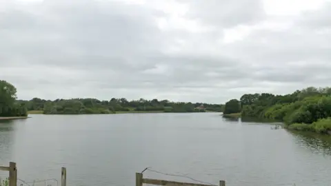 Google A stretch of water surrounded by trees and bushes on three sides and a road and fence on another.