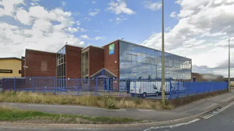 A general view picture of the depot building, which is made of brick and has a glass front. Three white vans are parked outside it.