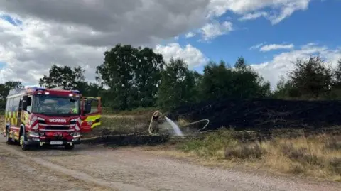 Staffordshire County Council A fire engine is parked on the road next to a scorched area of heath, as a firefighter sprays water on it. There are trees in the background.