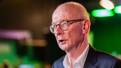 Getty Images A man in a navy blue suit with an open-necked white shirt collar. He is wearing glasses and balding. He appears to be giving an interview. There are green lights behind him but the background is blurred. He seems to be in a TV studio of some kind. 
