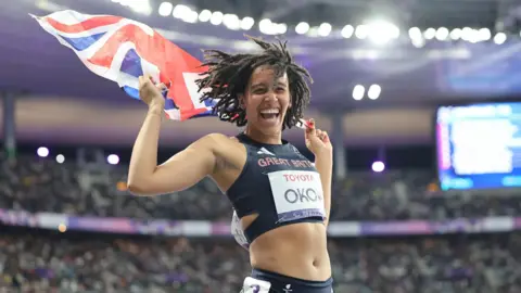 Getty Images Didi Okah - a woman with long brown hair and wearing a running kit - smiles widely as she celebrates on the track in a stadium while holding a union flag.