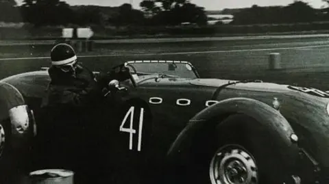 Sally Oliver The picture shows a vintage racing car on a track, captured in black and white. The car is an open-top sports racer with a sleek, aerodynamic body and prominent front wheel arches. It has the number 41 painted on the side. The driver is wearing protective gear, including a helmet and goggles, and is gripping the steering wheel while leaning into a turn.