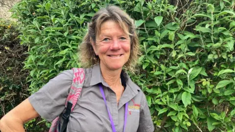 A woman with mid-length mousey blonde hair wearing a short sleeved green shirt and standing in front of a green leafy bush. There's a grey and pink coloured rucksack strap over her right shoulder and a purple lanyard hanging down the shirt.