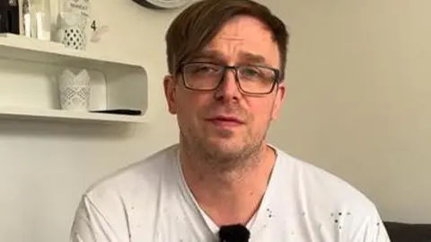 Gregory Lesiak sitting on a chair looking into a camera. He has dark hair and is wearing glasses and a white t-shirt.
