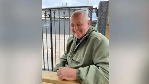 A man is sitting at a pub bench. He is wearing a green jacket and a blue top. He is resting his arm on the bench. He is bald and is smiling at the camera.