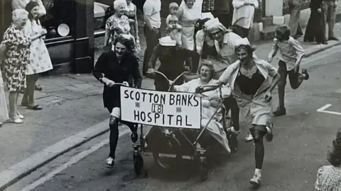 Six people running alonside a go-kart like bed with a lady sat on top for Scotton Banks Hospital