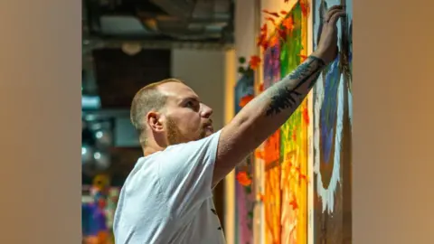 Gary Harper Gary Harper has short light brown hair and a short beard. He is wearing a white t-shirt and is reaching his arm up and touching a painting on a wall.