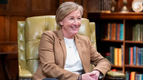 A close up shot of a middle-aged blonde woman sat in a leather chair in a room with a bookcase behind her smiling - Amanda Collier on The Traitors